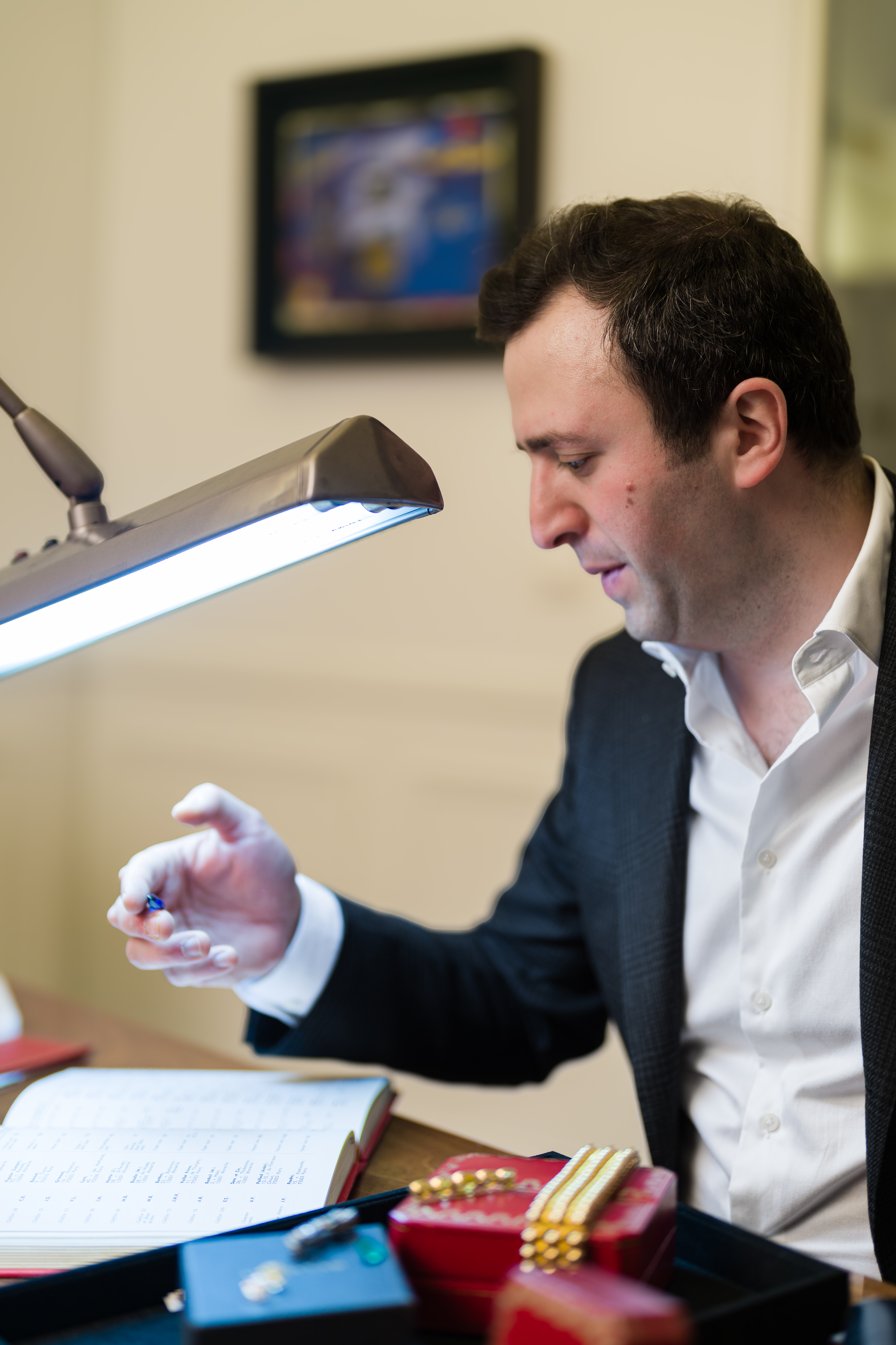 Ariel Altmann, négociant en bijoux à Paris, examinant une pierre précieuse sous la lampe d'expertise, entouré d'écrins et de bijoux signés